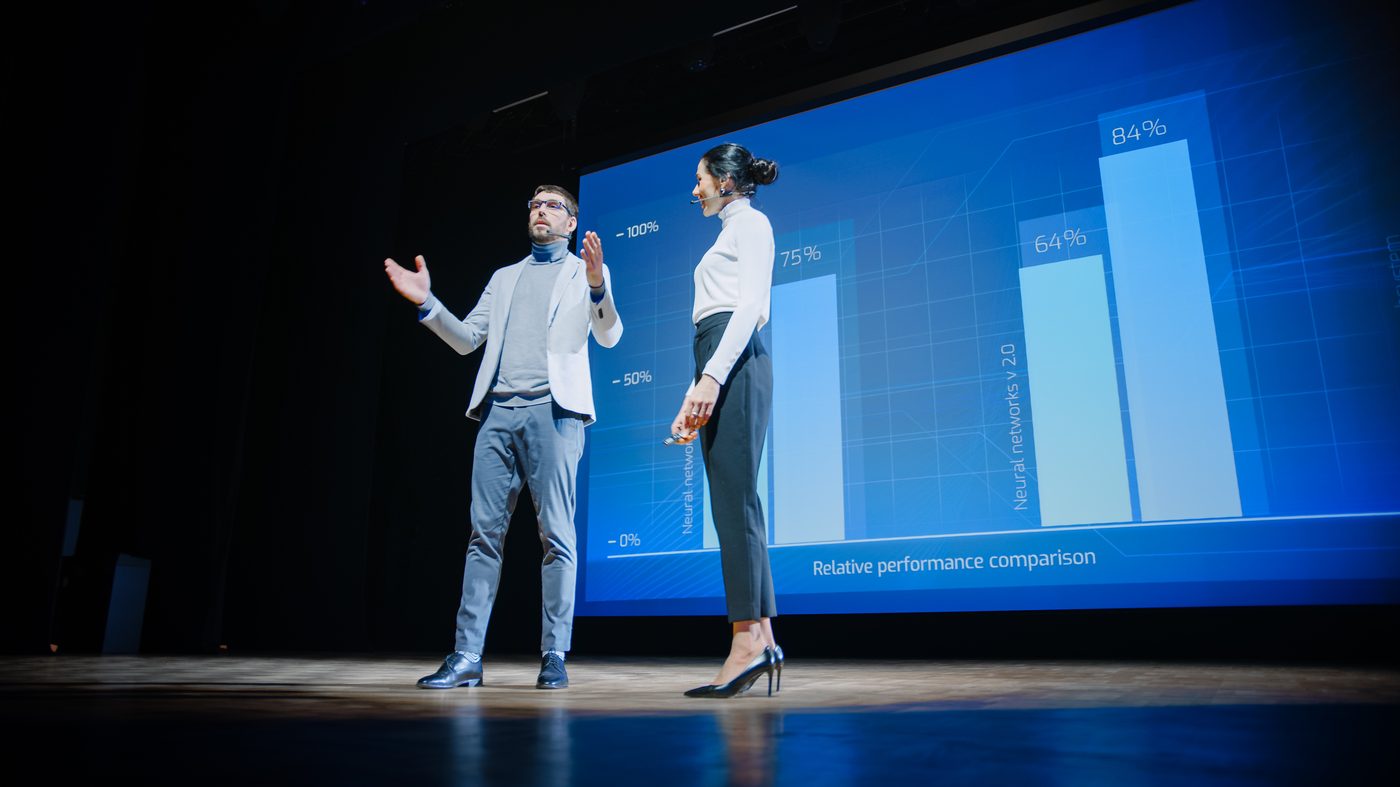 Two presenters on a keynote stage with a large chart display
